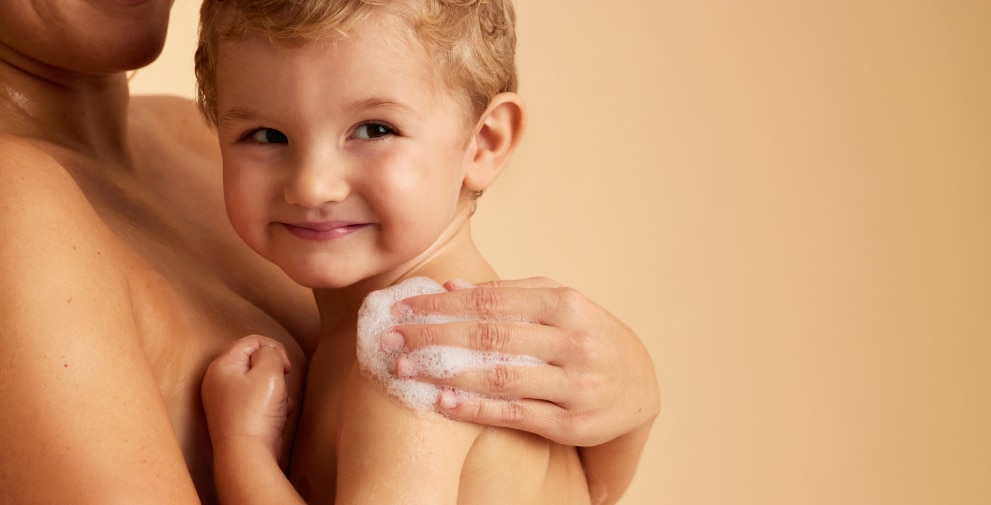 Een lachend, blond kindje kijkt in de camera terwijl een volwassene het vasthoudt en de schouder inzeept met schuim.