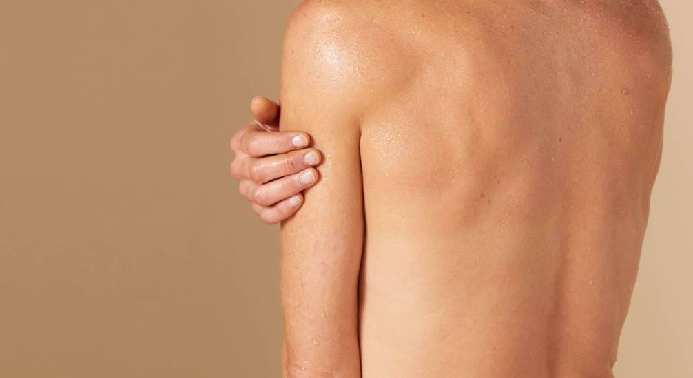 De achterkant van een persoon met een natte of glanzende huid, die de eigen bovenarm vasthoudt. De achtergrond is effen beige.