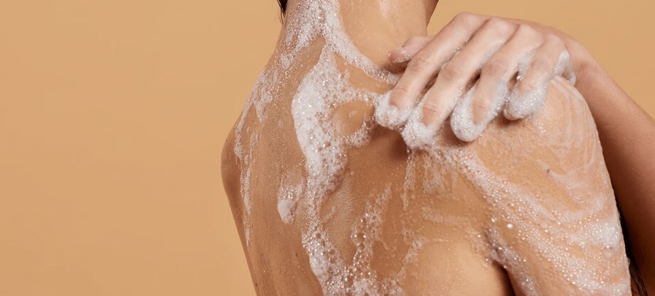 Achteraanzicht van een persoon die schuimende, parfumvrije douchegel aanbrengt op de schouder tegen een warme, beige achtergrond.
