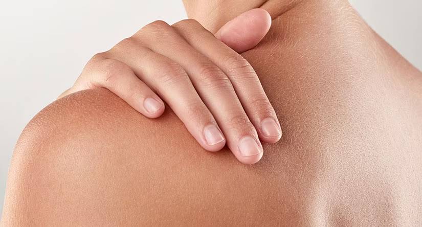 Close-up van een hand met verzorgde nagels die zachtjes op de eigen schouder rust. De achtergrond is lichtgrijs.