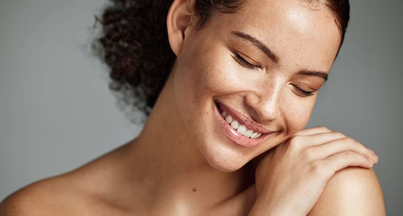 Close-up van een lachende vrouw met sproeten en opgestoken krullend haar. Ze heeft haar ogen gesloten en haar hand rust op haar schouder.