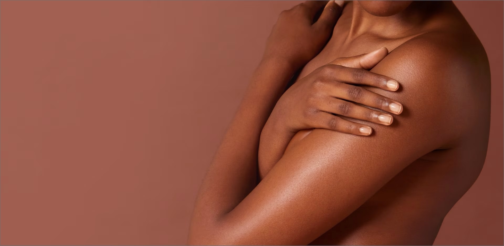 Close-up van een persoon met een donkere huid die zichzelf omarmt, waarbij de hand zachtjes op de schouder rust. De achtergrond is effen bruin.