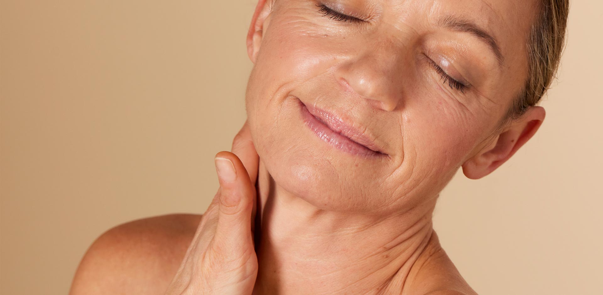 Close-up van een oudere vrouw met een rijpe huid die met gesloten ogen glimlacht. Ze raakt zachtjes haar nek aan, met een serene uitdrukking.