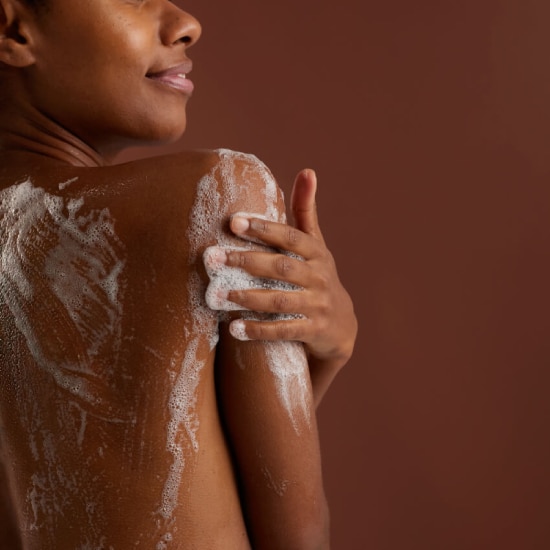 Close-up van de rug en schouder van een persoon met een donkere huid, die zichzelf inzeept met wit schuim tegen een bruine achtergrond.