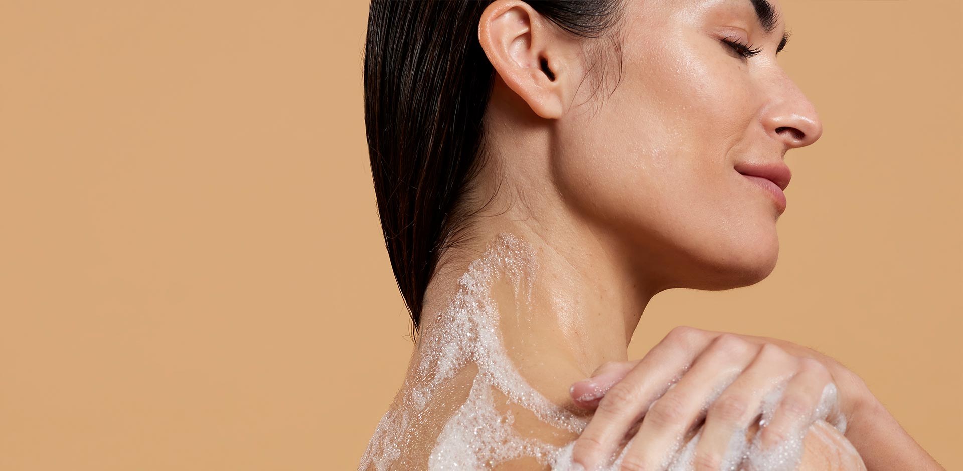 Een vrouw in profiel met gesloten ogen, die haar schouder wast met een rijk schuim tegen een beige achtergrond.