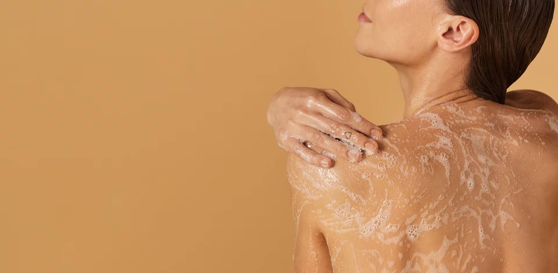 Een vrouw, gezien op haar rug, die haar schouder wast met een rijk schuim. Haar hand rust op de ingezeepte huid en de achtergrond is effen beige.an.