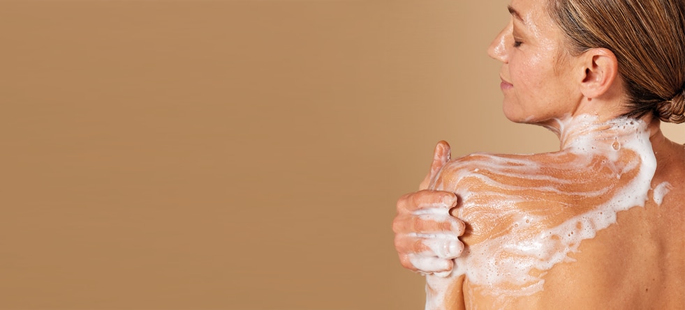 Een vrouw met een lichte huidskleur, gezien vanaf haar rug, die haar schouder inzeept met een rijk schuim tegen een beige achtergrond.