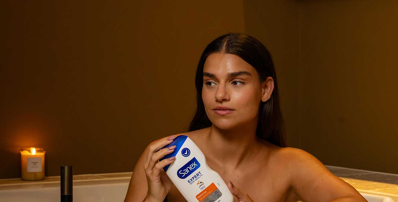 Een close-up van een ontspannen jonge vrouw die in een ligbad zit met een brandende kaars op de achtergrond, terwijl ze een flacon Sanex Expert Skin Health douchegel vasthoudt.